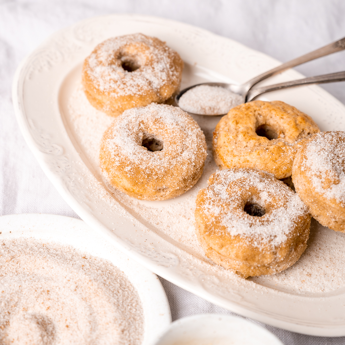 Baked cinnamon sugar donuts on a white decorative plate, with a spoonful and a bowl of cinnamon sugar nearby on a light textile surface.