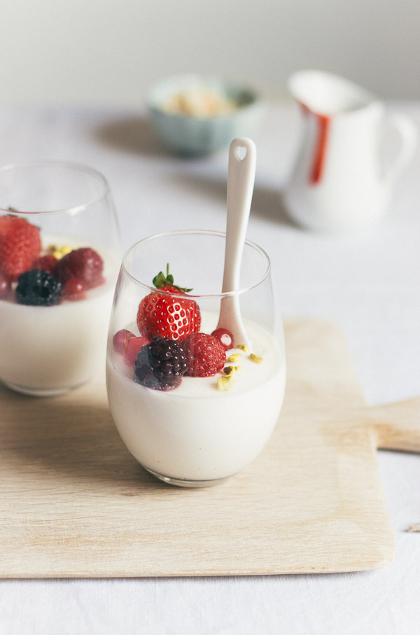 Image of classic panna cotta filled halfway up a stemless wine glass and topped with assorted berries and pistachio nuts with a heart-shaped porcelain spoon inserted.