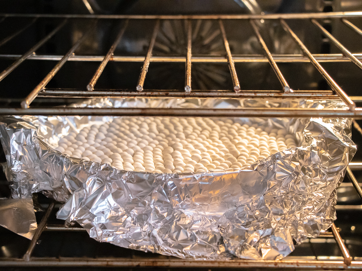 Quiche crust being blind baked in an oven with a layer of pie weights on top of the aluminum foil.