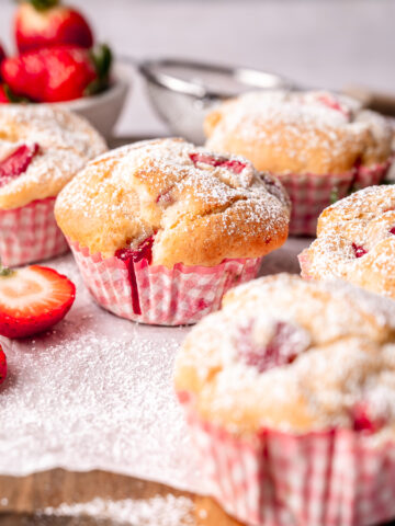 Sour Cream Strawberry muffins with strawberries peeking out of the domed tops covered in a dusting of powdered sugar with pink gingham cupcake liners.