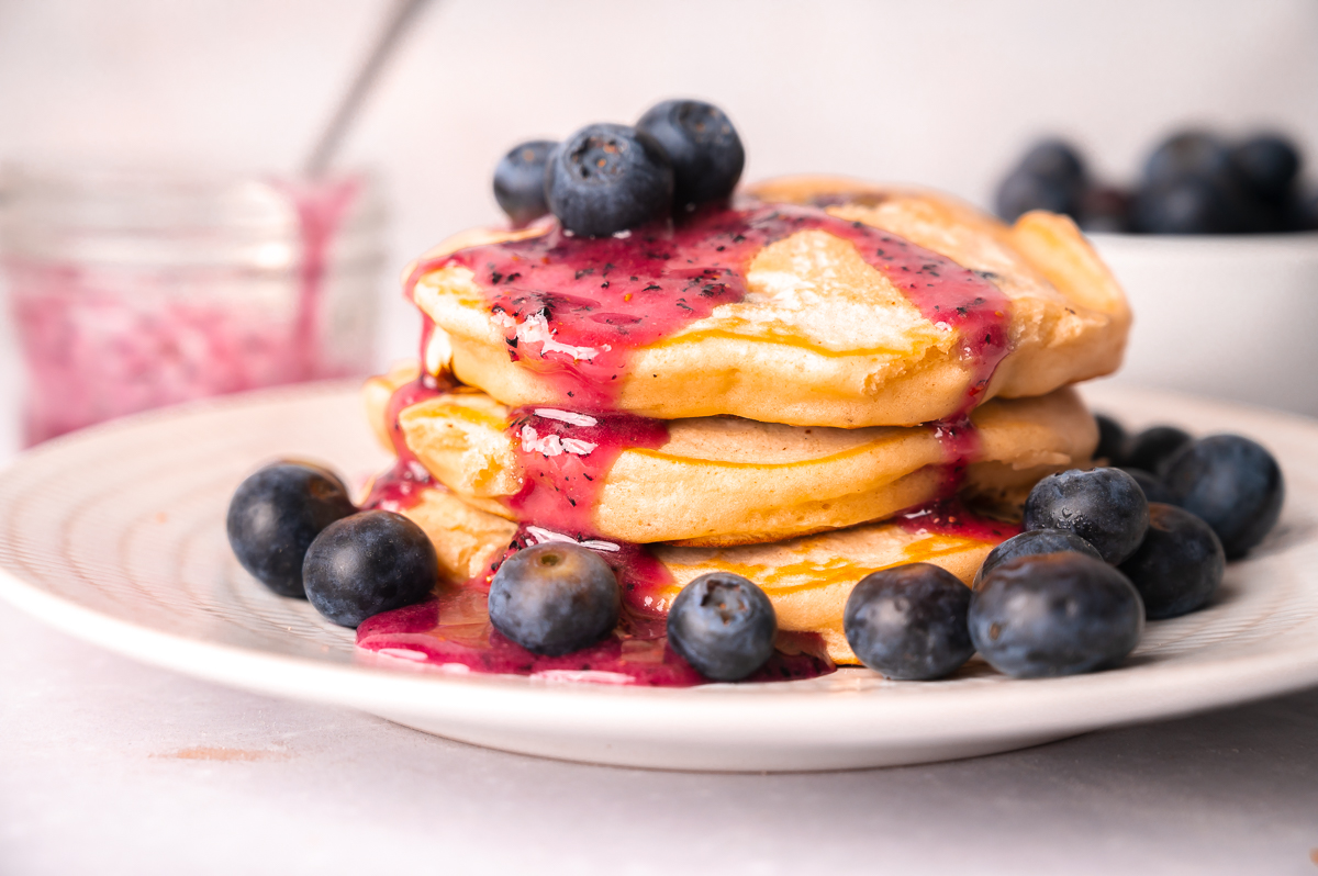 Stack of fluffy buttermilk blueberry pancakes with fresh homemade blueberry syrup and fresh berries on top.