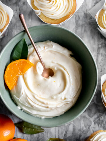 Orange Cream cheese frosting swirled in a bowl with an orange slice and frosted cupcakes on the side.