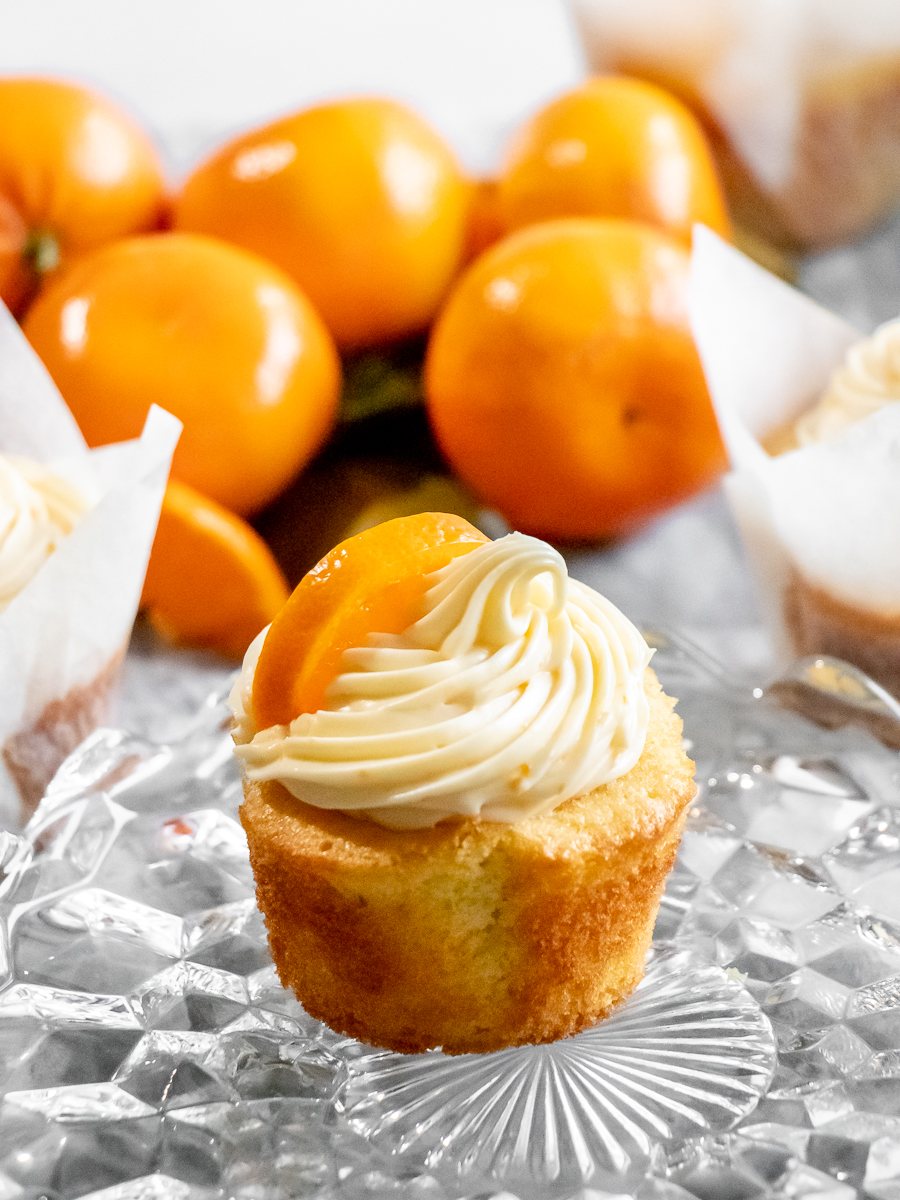 Citrusy orange cupcakes with a swirl of orange cream cheese frosting surrounded by fresh oranges.