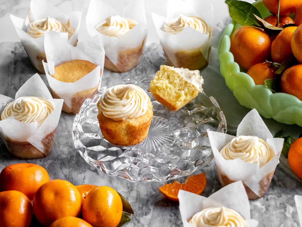Citrusy orange cupcakes, some cut in half, with orange cream cheese frosting surrounded by fresh oranges.