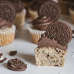 Cookies and cream cupcake split in half, topped with black cocoa cream cheese frosting and an oreo garnish.