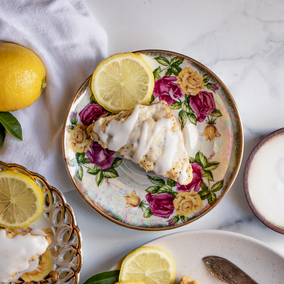 Close-up of a glazed lemon poppy seed scone served on a vintage floral plate with a slice of lemon.
