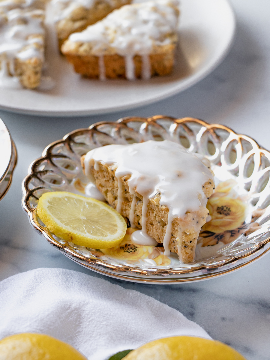 Close-up of a glazed lemon poppy seed scone served on a vintage floral plate with a slice of lemon.