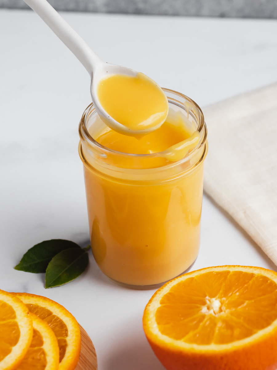 Scoop of orange curd being removed from a jar of orange curd, fresh oranges surround.