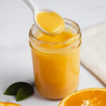 Creamy orange curd in a jar with a spoonful of curd being removed, flanked by fresh oranges.