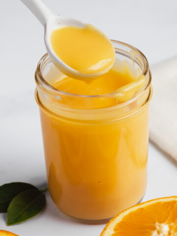 Creamy orange curd in a jar with a spoonful of curd being removed, flanked by fresh oranges.