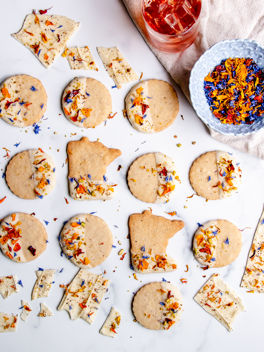 Minnesota shaped cookies dipped in white chocolate and decorated with dried flowers, pistachio, and edible gold flake.